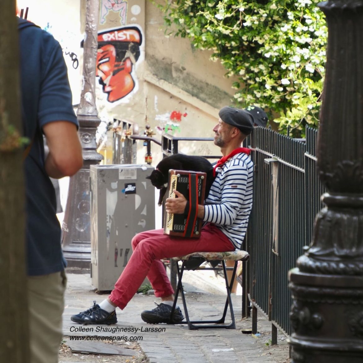 Classic scene of man wearing a beret, striped shirt, red scarf and trousers playing accordion with cat on top Place du Calvaire Montmartre Photo Walk May 2026